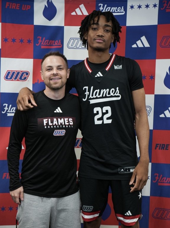 Solomon Bratton, wearing his UIC Flames jersey, poses with a UIC coach in front of a UIC backdrop.
